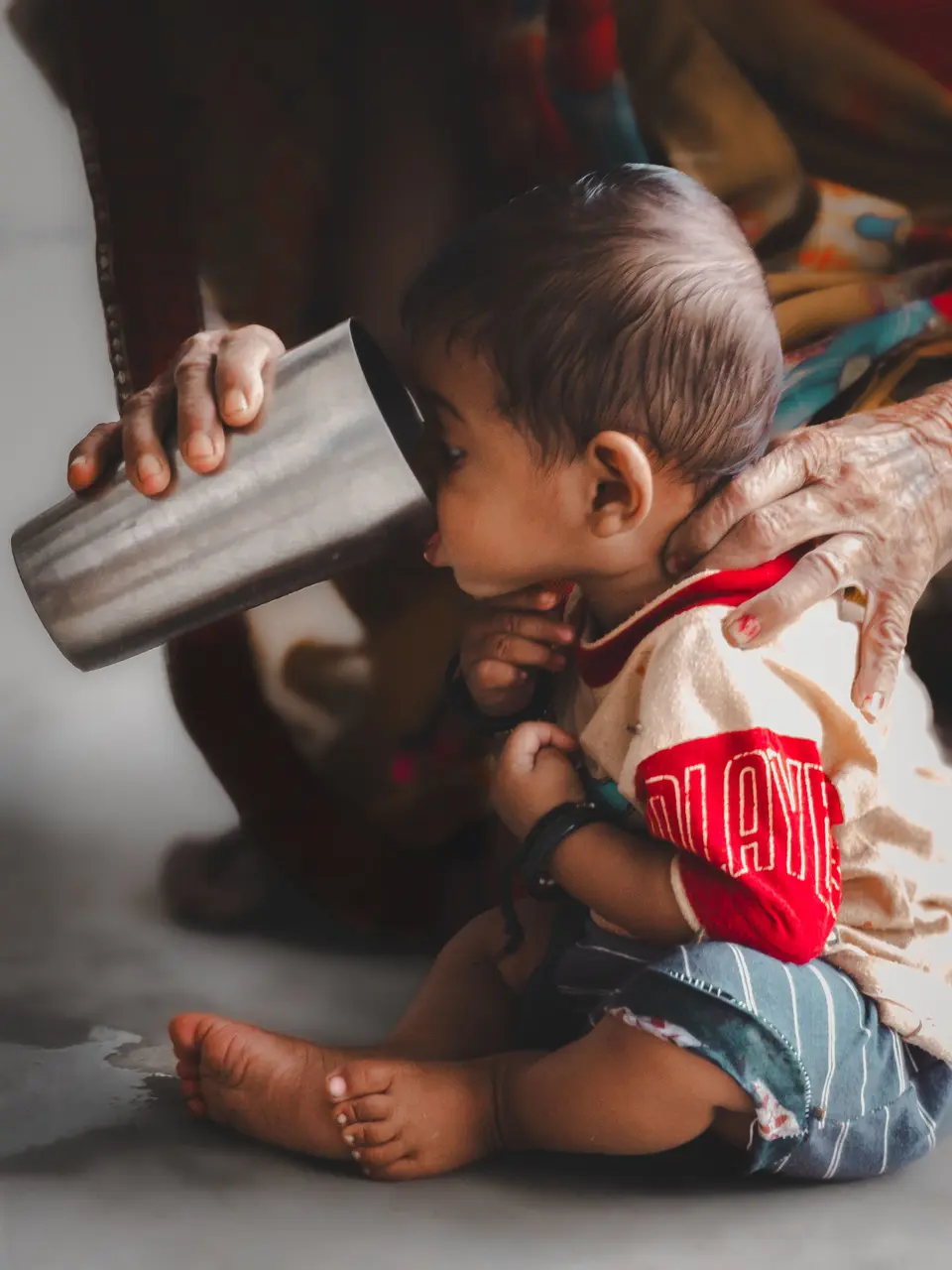 An Indian toddler sits on the floor drinking milk from a traditional steel tumbler held by an elder's hand.
