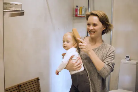 A mother smiles while gently brushing her baby's hair in a bright bathroom, both faces close together and clearly visible.