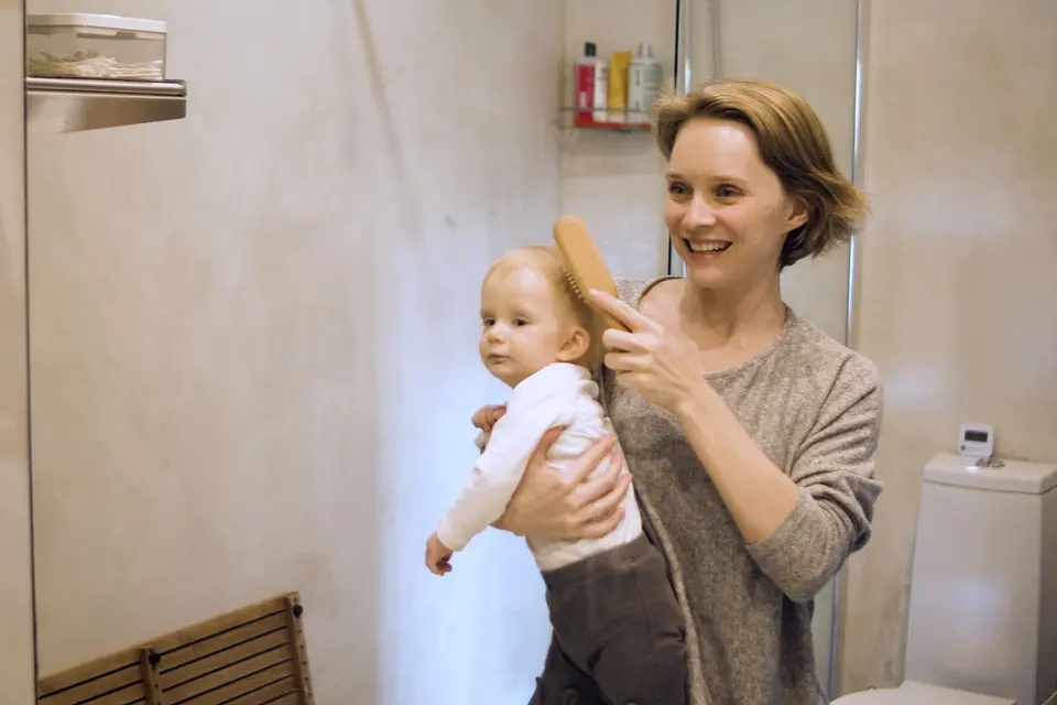 A mother smiles while gently brushing her baby's hair in a bright bathroom, both faces close together and clearly visible.