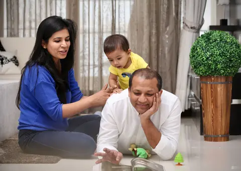 A mother and father sit on the floor with their toddler son, who leans over the father's shoulder, surrounded by toys and a picture book.