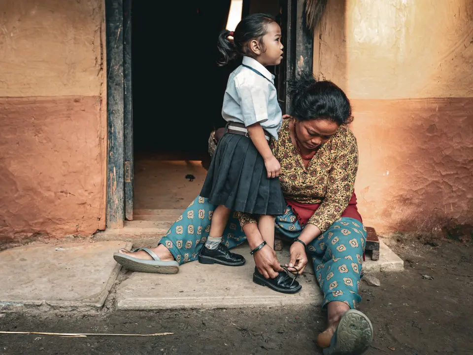A mother kneels to tie her young daughter's school shoes at a doorstep, both in South Asian dress, preparing for the school day.