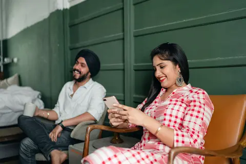 A smiling South Asian woman checks her phone in a cosy living room while her husband, in a turban, relaxes nearby with a cup of tea.