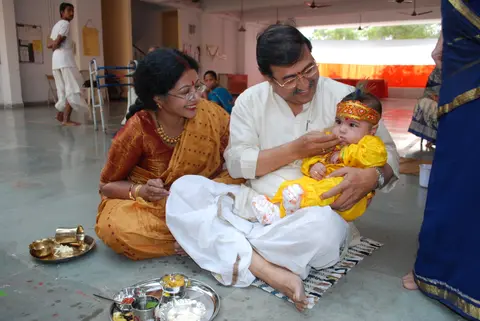 A father feeds his infant son, dressed in ceremonial yellow attire with a peacock-feather headband, during an Annaprashan first-rice-eating ceremony as the mother looks on.