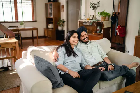 An Indian couple, the husband wearing a turban, sit close together on a sofa smiling.