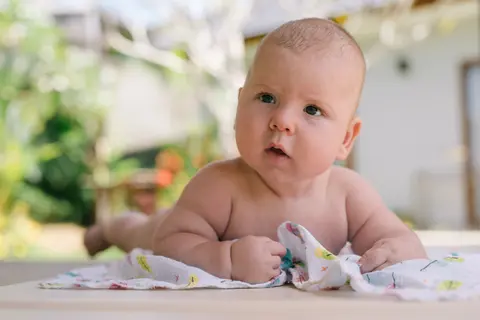 A baby doing tummy time outdoors, propped up on their arms, face and eyes clearly toward the camera, gripping a printed cloth.