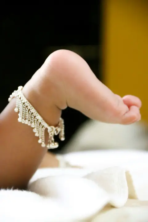 Close-up of a newborn's foot wearing a delicate silver anklet, resting on soft white fabric.
