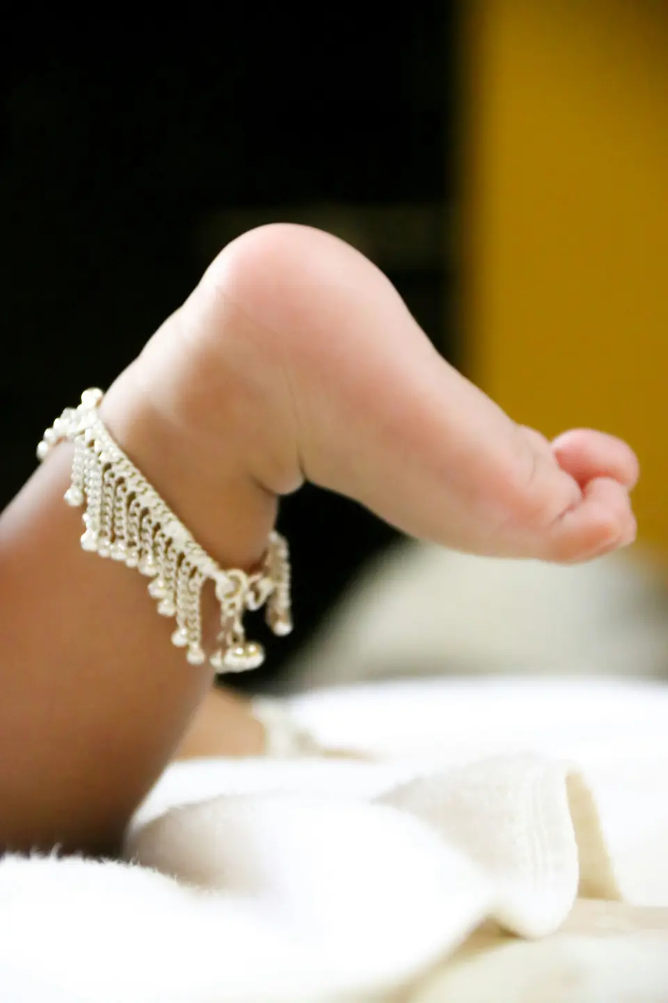 Close-up of a newborn's foot wearing a delicate silver anklet, resting on soft white fabric.