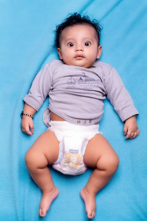 An Indian baby lies on his back on a blue blanket, alert and wide-eyed, wearing a grey onesie and diaper with legs relaxed and bare.