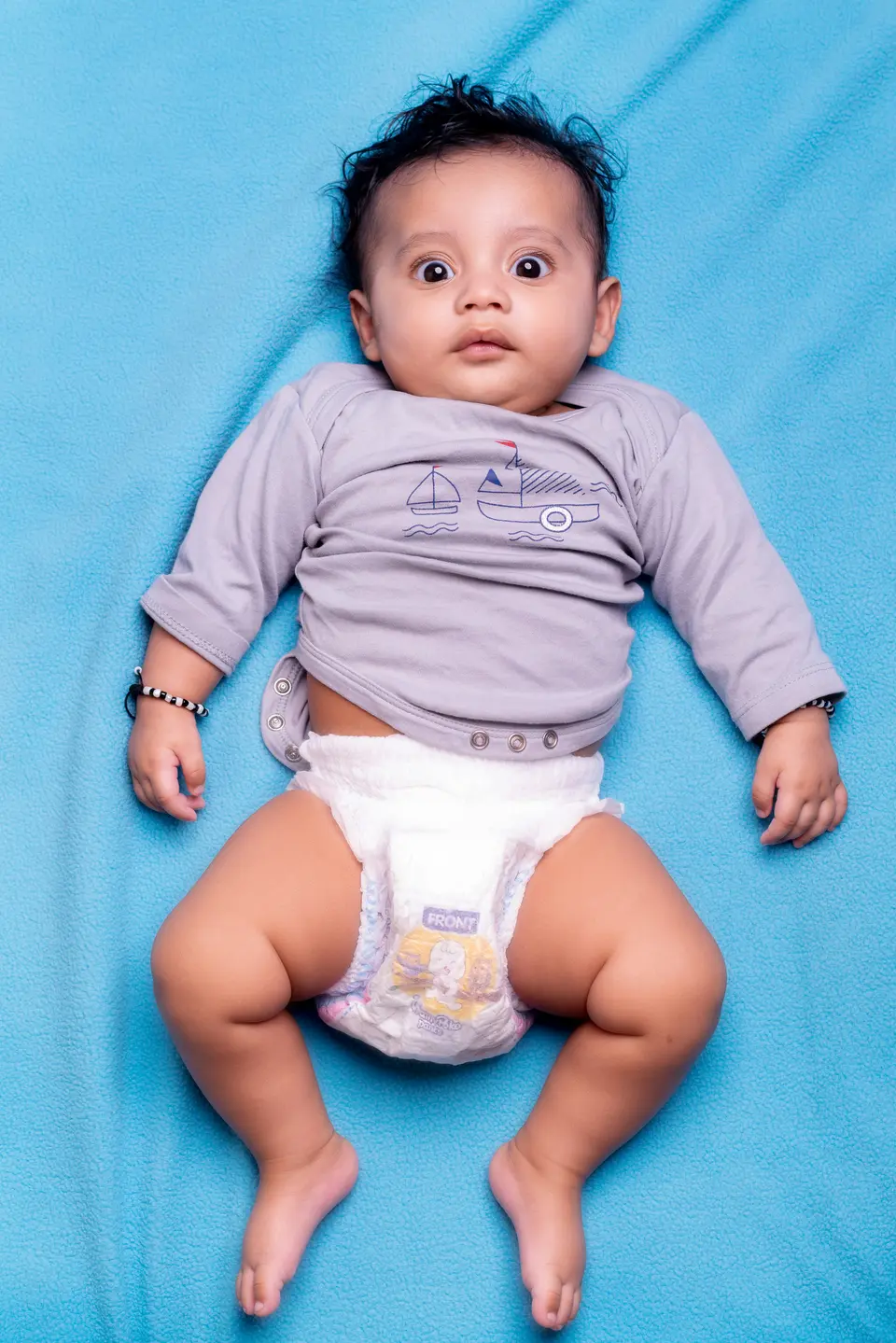 An Indian baby lies on his back on a blue blanket, alert and wide-eyed, wearing a grey onesie and diaper with legs relaxed and bare.