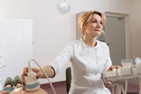 A doctor in a white coat performs an abdominal ultrasound scan on a patient lying on an exam table, no screen visible.