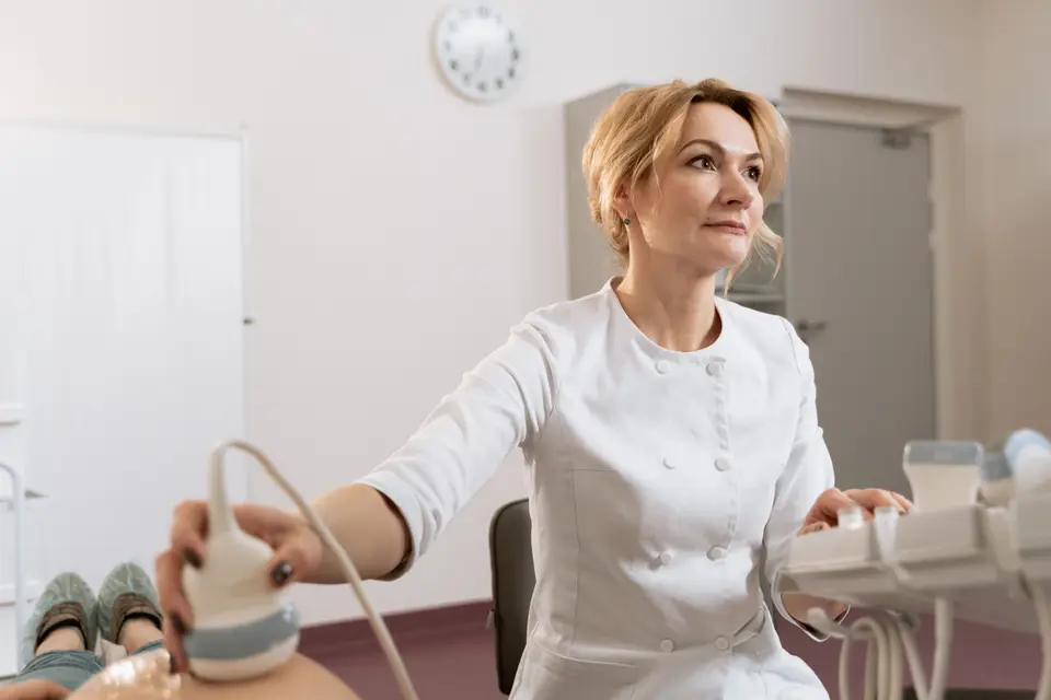A doctor in a white coat performs an abdominal ultrasound scan on a patient lying on an exam table, no screen visible.