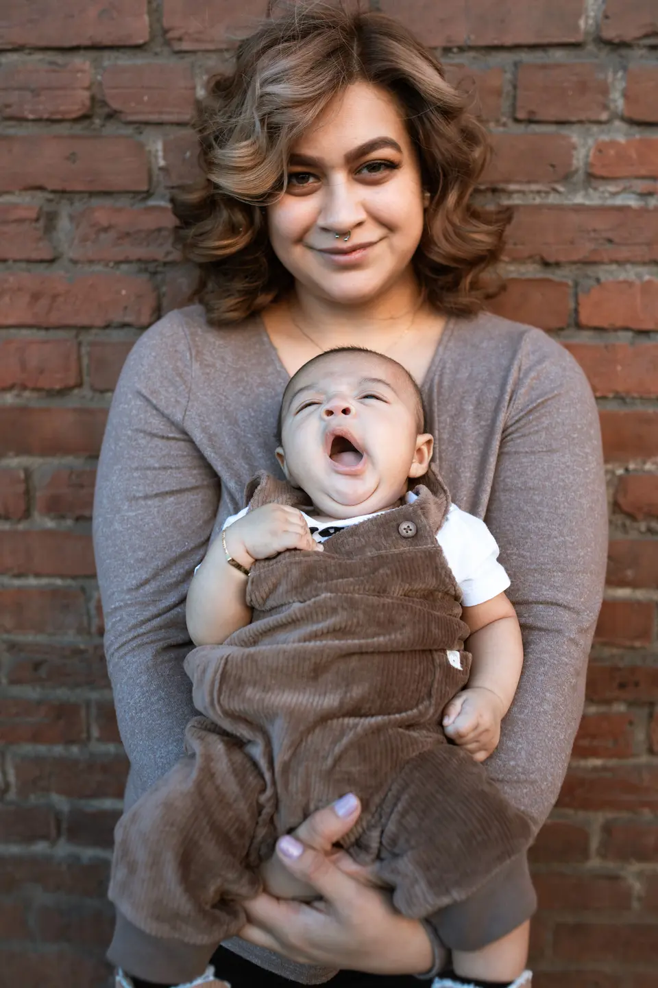 A mother holds her yawning baby upright against her shoulder outdoors in front of a brick wall, the baby's eyes closed mid-yawn.