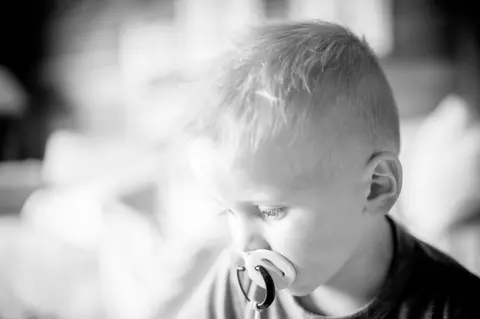 A close-up black-and-white portrait of a toddler boy with a pacifier in his mouth.