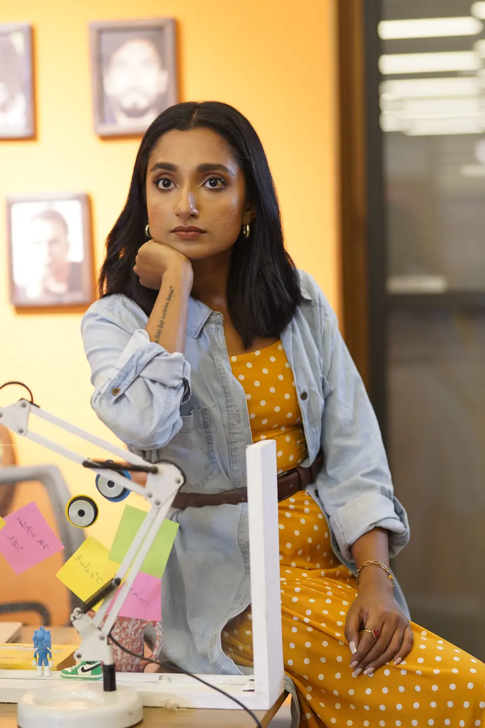 An Indian woman rests her chin on her hand at a desk in a creative office, with mood-board photos pinned to the wall behind her
